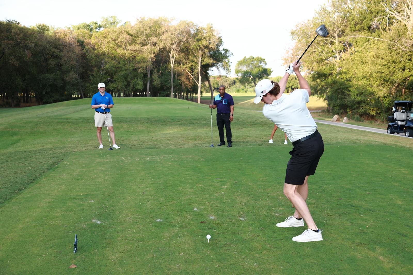A player at the top of the backswing on a tee box while two playing partners watch