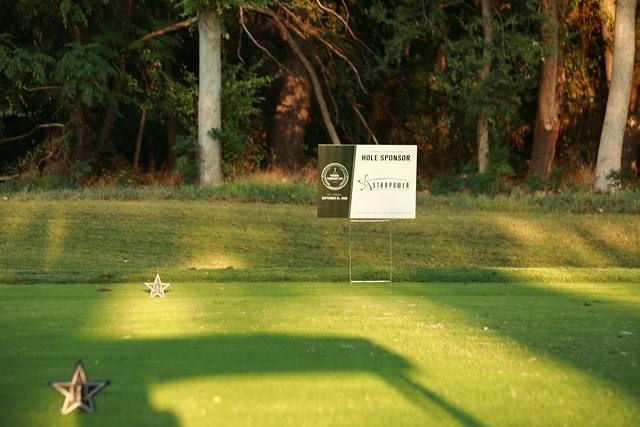 A hole sponsor sign and a Cowboys star tee marker in the morning sun