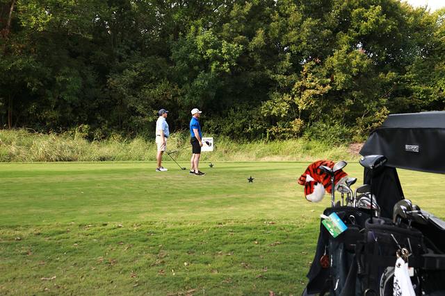 Two players on a tee box seen past a cart full of golf bags
