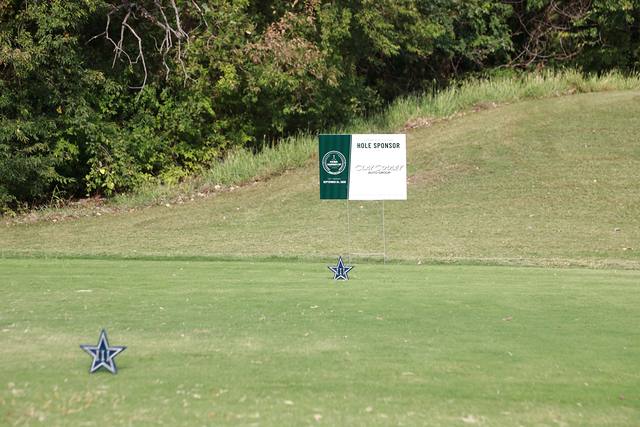 A hole sponsor sign on a tee box beside Cowboys star markers