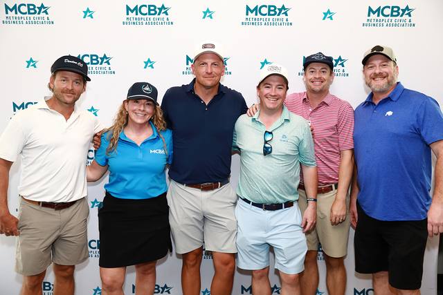 A team posing with an MCBA team member in front of the MCBA backdrop